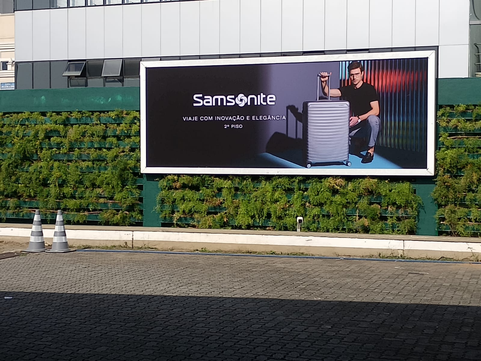 Painel publicitário Samsonite — lona externa sobre muro verde em shopping